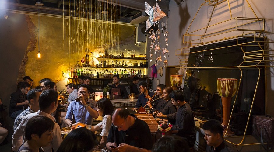 Moody interior of Tep Bar in Bangkok featuring an open bar counter, golden ambient lighting, and musicians playing traditional Thai instruments.